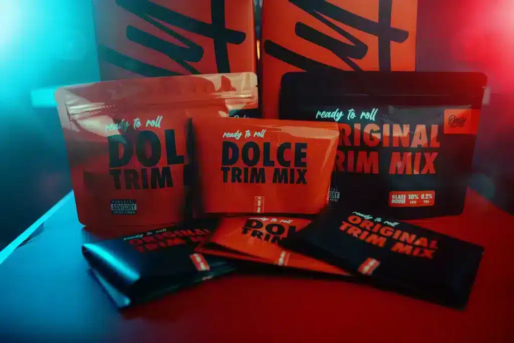La imagen presenta una disposición elegante de productos de CBD de la marca Only CBD, enfocándose en sus paquetes "DOLCE TRIM MIX" y "ORIGINAL TRIM MIX". Los paquetes, en tonos vibrantes de rojo y negro, están dispuestos sobre un fondo que complementa el esquema de color con grandes letras de la marca. La iluminación juega con sombras y reflejos, dando un aspecto sofisticado y de alta calidad a los productos, ideales para clientes conscientes de la marca y la estética en el nicho de productos de CBD.